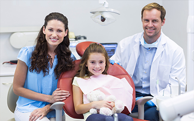 A young girl sitting in a dental chair holding a teddy bear with their mother and the dentist standing beside them